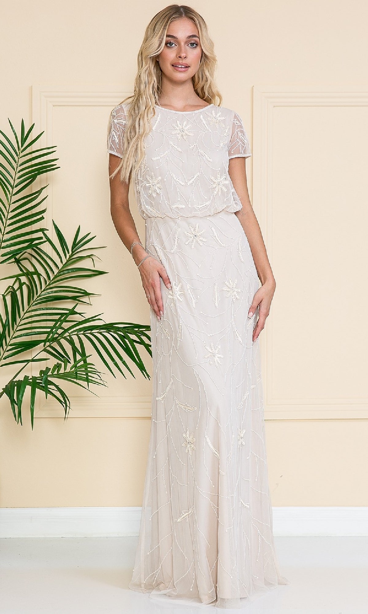A woman stands indoors in the Blouson Long Formal Dress: Amelia Couture IN004, a floor-length cream gown with short sleeves and floral embellishments. A leafy green plant sits on her left beside a beige wall.